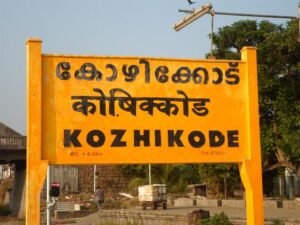 Yellow railway station signboard displaying "KOZHIKODE" in English, Malayalam, and Hindi, with trees and part of the railway station visible in the background under a clear sky. used in a blog of 'why you have to visit calicut at least once'.